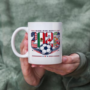 Puede incluir: Una taza de cer&aacute;mica blanca con el texto 2026, donde los n&uacute;meros est&aacute;n rellenos con las banderas de Estados Unidos, M&eacute;xico y Canad&aacute;, junto a un gr&aacute;fico de un bal&oacute;n de f&uacute;tbol y el texto ONE NATION - ONE GAME - ONE WORLD con estrellas.