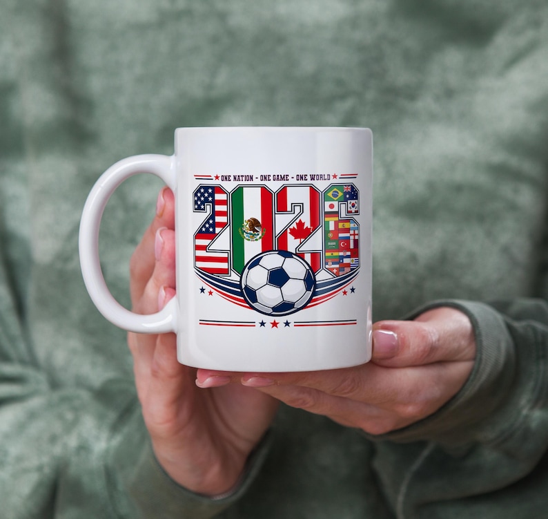 Puede incluir: Una taza de cer&aacute;mica blanca con el texto 2026, donde los n&uacute;meros est&aacute;n rellenos con las banderas de Estados Unidos, M&eacute;xico y Canad&aacute;, junto a un gr&aacute;fico de un bal&oacute;n de f&uacute;tbol y el texto ONE NATION - ONE GAME - ONE WORLD con estrellas.