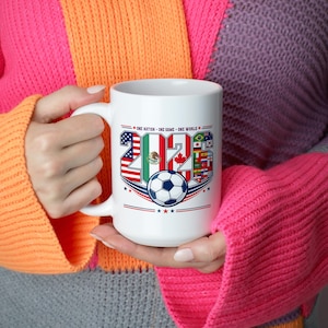 Puede incluir: Una taza de cer&aacute;mica blanca con un dise&ntilde;o gr&aacute;fico que incluye el texto 'ONE NATION - ONE GAME - ONE WORLD' y el a&ntilde;o '2026'. Los n&uacute;meros est&aacute;n rellenos con varias banderas nacionales y un bal&oacute;n de f&uacute;tbol blanco y negro est&aacute; centrado bajo el texto.