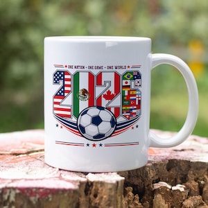 Puede incluir: Una taza de cer&aacute;mica blanca con el texto 'ONE NATION - ONE GAME - ONE WORLD' sobre el a&ntilde;o '2026'. Los n&uacute;meros est&aacute;n rellenos con varias banderas nacionales, incluyendo las de EE. UU., M&eacute;xico y Canad&aacute;, y un bal&oacute;n de f&uacute;tbol centrado debajo.