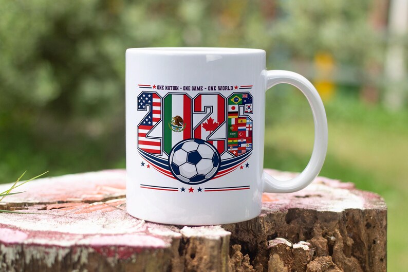 Puede incluir: Una taza de cer&aacute;mica blanca con el texto 'ONE NATION - ONE GAME - ONE WORLD' sobre el a&ntilde;o '2026'. Los n&uacute;meros est&aacute;n rellenos con varias banderas nacionales, incluyendo las de EE. UU., M&eacute;xico y Canad&aacute;, y un bal&oacute;n de f&uacute;tbol centrado debajo.
