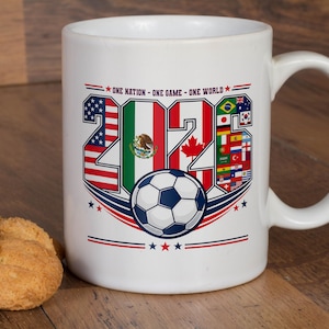 Puede incluir: Una taza de cer&aacute;mica blanca con un dise&ntilde;o gr&aacute;fico que incluye el texto "ONE NATION - ONE GAME - ONE WORLD" sobre el a&ntilde;o "2026". Los n&uacute;meros est&aacute;n rellenos con varias banderas nacionales y un bal&oacute;n de f&uacute;tbol est&aacute; centrado bajo el texto.