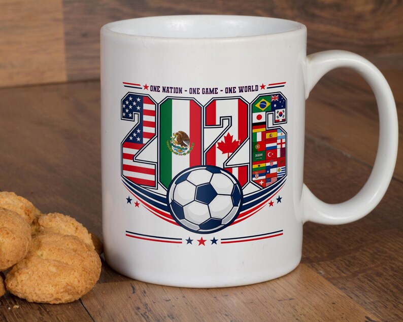Puede incluir: Una taza de cer&aacute;mica blanca con un dise&ntilde;o gr&aacute;fico que incluye el texto "ONE NATION - ONE GAME - ONE WORLD" sobre el a&ntilde;o "2026". Los n&uacute;meros est&aacute;n rellenos con varias banderas nacionales y un bal&oacute;n de f&uacute;tbol est&aacute; centrado bajo el texto.