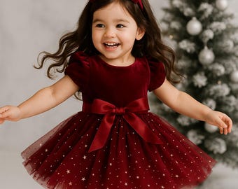 May include: A burgundy velvet dress with a tulle overlay scattered with gold stars. The dress features a matching bow at the waist and a headband. The outfit is completed with burgundy Mary Jane shoes.