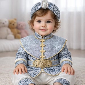 May include: A baby wearing a blue and white patterned outfit, including a turban with a jewel, a jacket with gold buttons, and white pants. The outfit has gold trim and a decorative gold belt. The baby is sitting on a white rug.