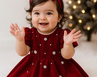 May include: A smiling baby wearing a red velvet dress with pearl accents and a matching bow headband. The dress has a flared skirt and short sleeves. The baby is also wearing red shoes. A pearl necklace is around the baby's neck.