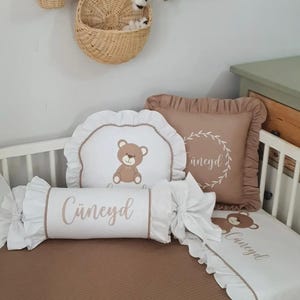 May include: A baby bedding set in brown and white, featuring a teddy bear design and the name "Cüneyd". The set includes a fitted sheet, a pillow, and a bolster pillow with ruffled edges. A woven basket hangs on the wall.