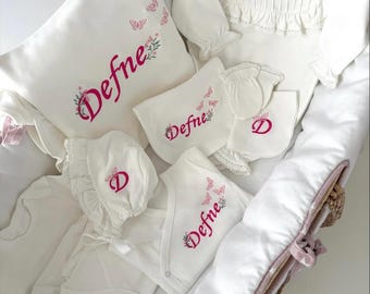 May include: A collection of white baby clothing and accessories, including a pillow, blanket, and onesie, all embroidered with the name "Defne" in pink. The set features floral and butterfly motifs.