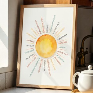 May include: A framed print with a watercolor sun design and affirmations, displayed on a kitchen countertop. The print features phrases like "I am calm" and "I am worthy." A white teapot and a wooden plate are also on the counter.