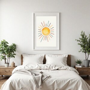 May include: A framed art print featuring a watercolor sun design with positive affirmations in a circular pattern. The artwork is in a white frame and hangs above a bed with white bedding. The affirmations include phrases like "I am brave" and "I am strong."