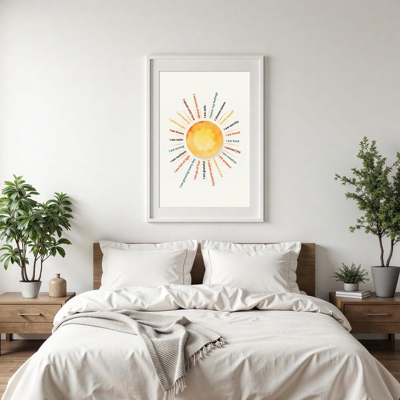 May include: A framed art print featuring a watercolor sun design with positive affirmations in a circular pattern. The artwork is in a white frame and hangs above a bed with white bedding. The affirmations include phrases like "I am brave" and "I am strong."