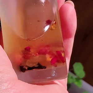 May include: A clear, rectangular bottle filled with a light amber liquid and small red flower petals. The bottle is held in a hand, with a blurred background of green plants.