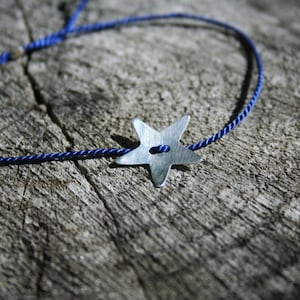 May include: A blue cord bracelet with a silver star charm. The star has a small button in the center.