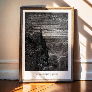 Könnte beinhalten: Gerahmtes Schwarz-Weiß-Kunstwerk mit dem Titel "SATAN CONTEMPLE L'EDEN", das eine detaillierte Illustration einer Figur auf einer Klippe mit Blick auf eine Landschaft zeigt. Das Kunstwerk befindet sich in einem Holzrahmen und wird vor einer neutralen Wand ausgestellt.