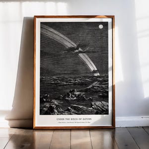 Op de afbeelding: Een ingelijste zwart-wit print getiteld Under the Rings of Saturn, met een hemels landschap met een grote planetaire ring die over een rotsachtig, bekraterd oppervlak boogt en een kleine maan in de donkere lucht.