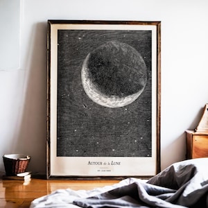 Puede incluir: Una impresión en blanco y negro enmarcada que presenta una ilustración detallada de la luna sobre un fondo estrellado. La impresión incluye el texto "AUTOUR de la Lune" y "JULES VERNE". La obra de arte se presenta en un marco de madera.