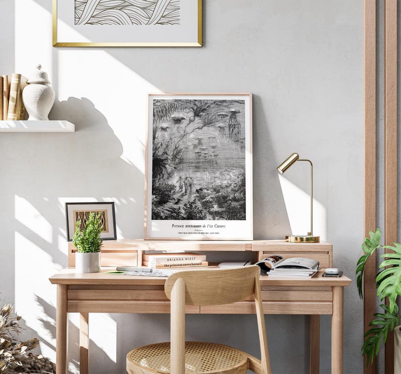 May include: A light wood desk with a framed print of a black and white illustration. The print has the text "Prince Roumanie & Ile Crepo". A small potted plant, books, and a gold desk lamp are also on the desk.