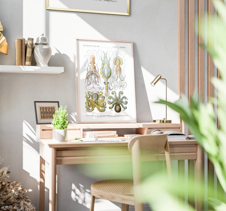 May include: A framed vintage-style print of various octopus species in a light wood frame. The print is displayed on a light wood desk with a small gold desk lamp. The desk is in a well-lit room with a neutral color palette.