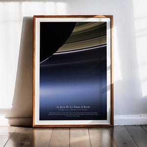 May include: A framed print depicting Saturn and its rings, with a colour gradient of blue and black. The print has French text and is in a wooden frame, leaning against a white wall. The print is approximately 60 cm x 80 cm.
