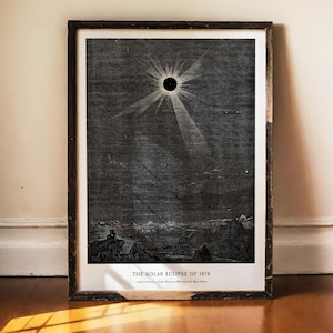 Puede incluir: Una impresión enmarcada en blanco y negro que representa el eclipse solar de 1878. La obra muestra un cielo oscuro con una corona solar radiante sobre un paisaje montañoso. El texto en la parte inferior dice THE SOLAR ECLIPSE OF 1878, Popular Astronomy, Camille Flammarion, 1880, Engraved by Rapine-Behrens.