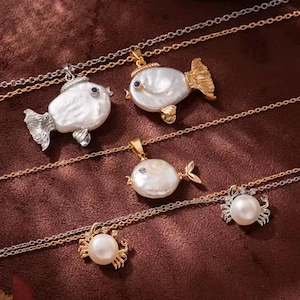 Puede incluir: Una colección de cinco collares con colgantes en forma de criaturas marinas. Tres colgantes son peces de perla barroca con aletas metálicas y ojos de piedra azul o clara. Dos colgantes son cangrejos hechos de perlas redondas.