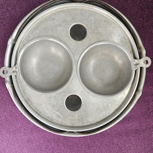 May include: A vintage aluminum egg poacher with two circular cups and a central hole. The poacher has a handle on each side and a circular rim. The poacher sits on a purple surface.