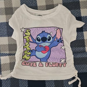 May include: A white short-sleeved t-shirt featuring a graphic of the blue character Stitch holding a red heart. The design includes the text "STITCH" and "CUTE & FLUFFY" with a purple checkered background and white stars. The sides have adjustable drawstrings.