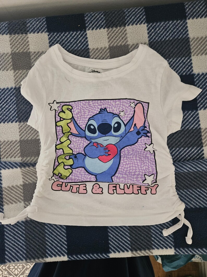 May include: A white short-sleeved t-shirt featuring a graphic of the blue character Stitch holding a red heart. The design includes the text "STITCH" and "CUTE & FLUFFY" with a purple checkered background and white stars. The sides have adjustable drawstrings.