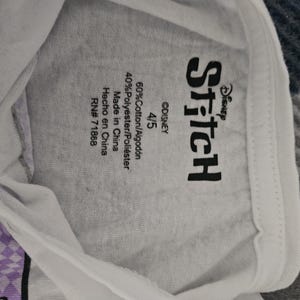May include: A white t-shirt featuring a black Disney Stitch logo printed on the inside neck area. The label includes the size 4/5, material composition of 60% cotton and 40% polyester, origin as Made in China, and the RN number 71868.