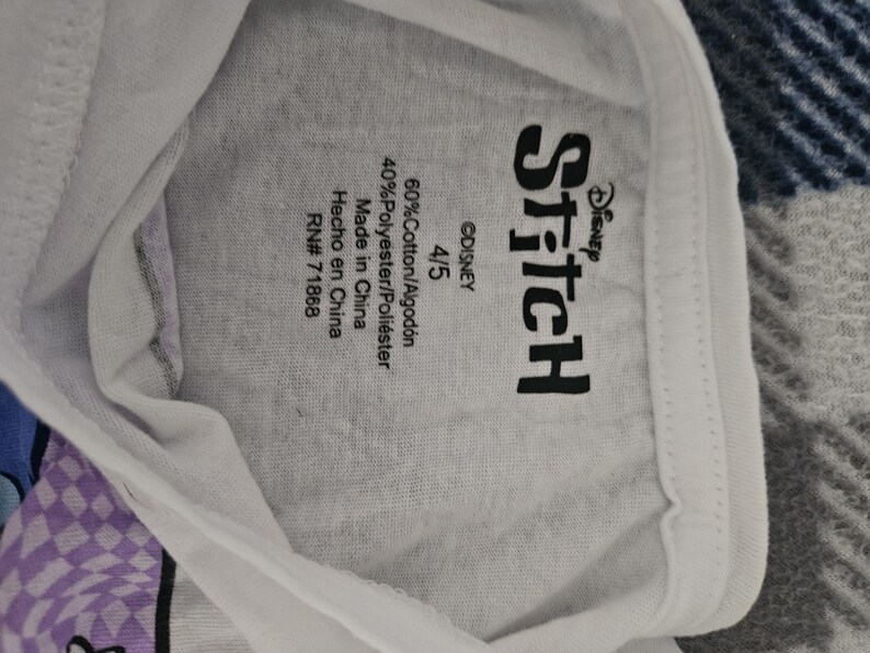 May include: A white t-shirt featuring a black Disney Stitch logo printed on the inside neck area. The label includes the size 4/5, material composition of 60% cotton and 40% polyester, origin as Made in China, and the RN number 71868.