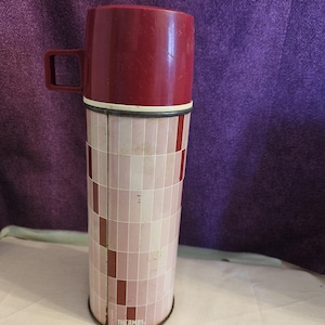 May include: A cylindrical Thermos brand vacuum flask featuring a maroon plastic cup lid with a side handle. The body is decorated with a grid pattern of light pink, white, and dark maroon rectangular tiles. The word THERMOS is printed at the base.