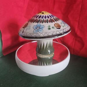 May include: A ceramic mushroom-shaped decorative piece with a white base and a red interior. The mushroom cap is adorned with blue flowers, butterflies, and other colorful designs. The stem is green with a textured pattern.