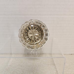 May include: A clear, faceted glass doorknob with a circular design. The knob has a detailed, textured surface and a central starburst pattern. The edges are scalloped, adding to its vintage aesthetic. The doorknob is displayed on a clear acrylic surface.
