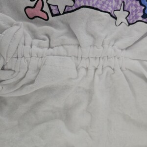 May include: A white garment featuring a central vertical drawstring channel with gathered fabric. The top portion displays a graphic print with purple checkered patterns, pink shapes, blue elements, and white star outlines on a white background.