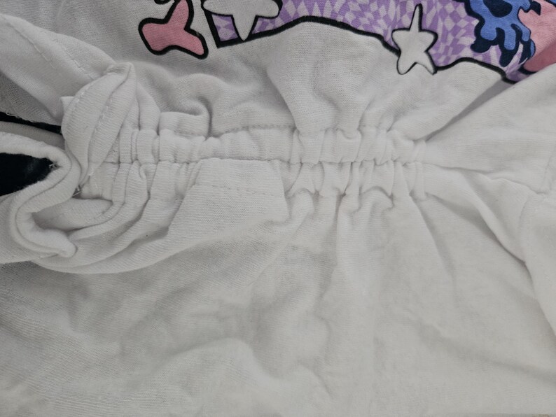 May include: A white garment featuring a central vertical drawstring channel with gathered fabric. The top portion displays a graphic print with purple checkered patterns, pink shapes, blue elements, and white star outlines on a white background.