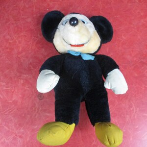 Op de afbeelding: Een vintage Mickey Mouse pluche speelgoed. Het pluche heeft een zwarte body, grote zwarte oren, een cr&egrave;mekleurig gezicht, witte handschoenen en gele schoenen. Een blauwe vlinderdas voegt een vleugje kleur toe. Een klassiek verzamelobject voor kinderen.