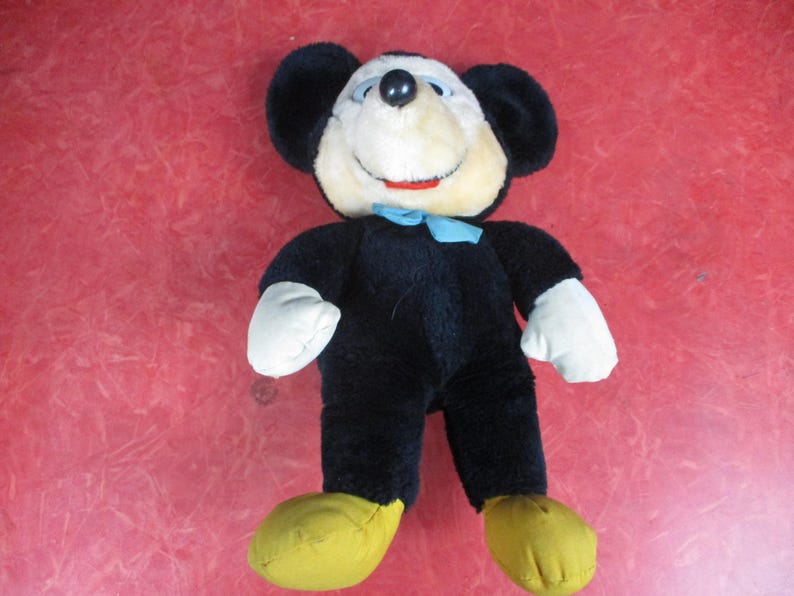 Op de afbeelding: Een vintage Mickey Mouse pluche speelgoed. Het pluche heeft een zwarte body, grote zwarte oren, een cr&egrave;mekleurig gezicht, witte handschoenen en gele schoenen. Een blauwe vlinderdas voegt een vleugje kleur toe. Een klassiek verzamelobject voor kinderen.