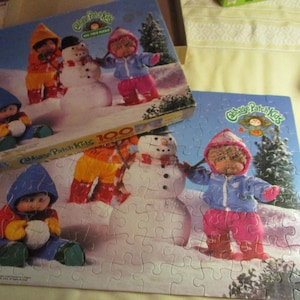 May include: A 100-piece Cabbage Patch Kids jigsaw puzzle featuring a winter scene with snowmen and dolls in winter attire. The puzzle box is visible, displaying the MB logo and the puzzle's theme.
