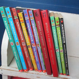 Puede incluir: Una colección de libros de Animorphs con cubiertas coloridas, incluyendo títulos como "The Stranger" y "The Alien". Los libros están colocados en una estantería blanca, con los lomos a la vista.