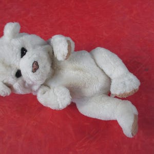 May include: A white plush dog toy with black eyes and a pink nose lies on a red surface. The dog's ears and paws have tan accents. The toy is soft and fluffy, suggesting a cuddly design.