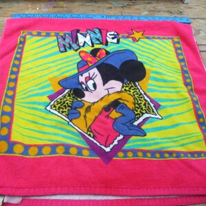 May include: A pink Minnie Mouse towel featuring a colorful design. The towel has a bright pink border with a yellow and green center. The Minnie Mouse graphic is wearing a blue hat and a yellow scarf. The word "MINNIE" is across the top.