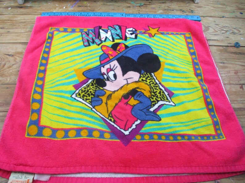 May include: A pink Minnie Mouse towel featuring a colorful design. The towel has a bright pink border with a yellow and green center. The Minnie Mouse graphic is wearing a blue hat and a yellow scarf. The word "MINNIE" is across the top.