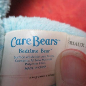 May include: A close-up of a tag from a Care Bears Bedtime Bear plush toy. The tag is white with blue text that reads "Care Bears Bedtime Bear". The tag also includes washing instructions and material information.