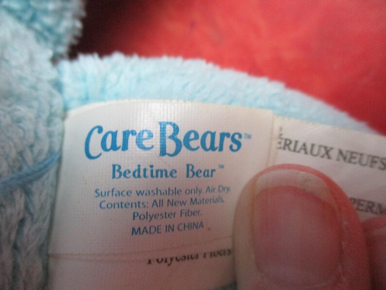 May include: A close-up of a tag from a Care Bears Bedtime Bear plush toy. The tag is white with blue text that reads "Care Bears Bedtime Bear". The tag also includes washing instructions and material information.
