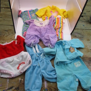 May include: A collection of doll clothing including a red and white romper with Cabbage Patch Kids text, a purple outfit with a lace collar, a blue hooded jumpsuit, a light blue corduroy set with a bear patch, and a yellow top with striped pants.