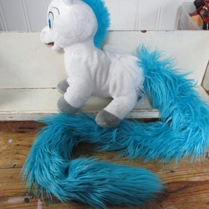 May include: A white plush toy of a mythical creature with a bright blue mane and a long, flowing blue faux fur tail. The creature has gray hooves and large blue eyes. A blue measuring tape is visible, showing a length of approximately 16 inches.