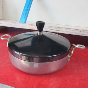 May include: A vintage metal cooking pot with a black lid and a black handle. The pot has two silver handles and a silver base. The pot is sitting on a red surface.