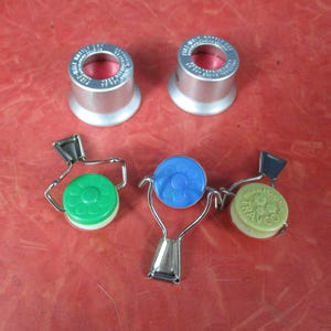May include: An assortment of bottle stoppers and caps. Two silver-colored metal stoppers with red interiors are at the top. Below are three bottle caps in green, blue, and light green, each with a metal wire closure.
