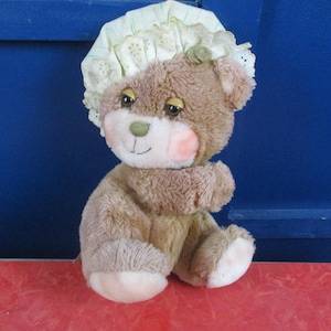 Puede incluir: Un oso de peluche marrón con un gorro verde claro con ribete de encaje. El oso tiene una nariz y mejillas rosadas, con patas color crema y una sonrisa cosida. El oso está sentado sobre una superficie roja contra un fondo azul.