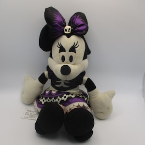Puede incluir: Mu&ntilde;eca de peluche de Minnie Mouse con disfraz de Halloween. La mu&ntilde;eca tiene una combinaci&oacute;n de colores en blanco y negro con un lazo y una falda morados. El lazo presenta una calavera. La mu&ntilde;eca tiene zapatos y guantes negros.